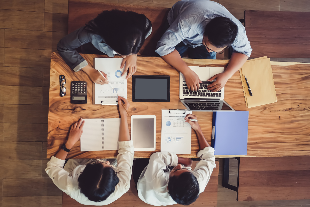 Four people reviewing financial reports, tablets and a laptop around a shared table