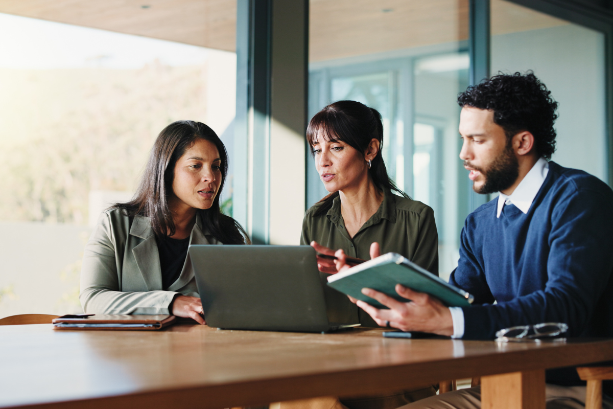 Three colleagues in a focused discussion around a laptop and notes